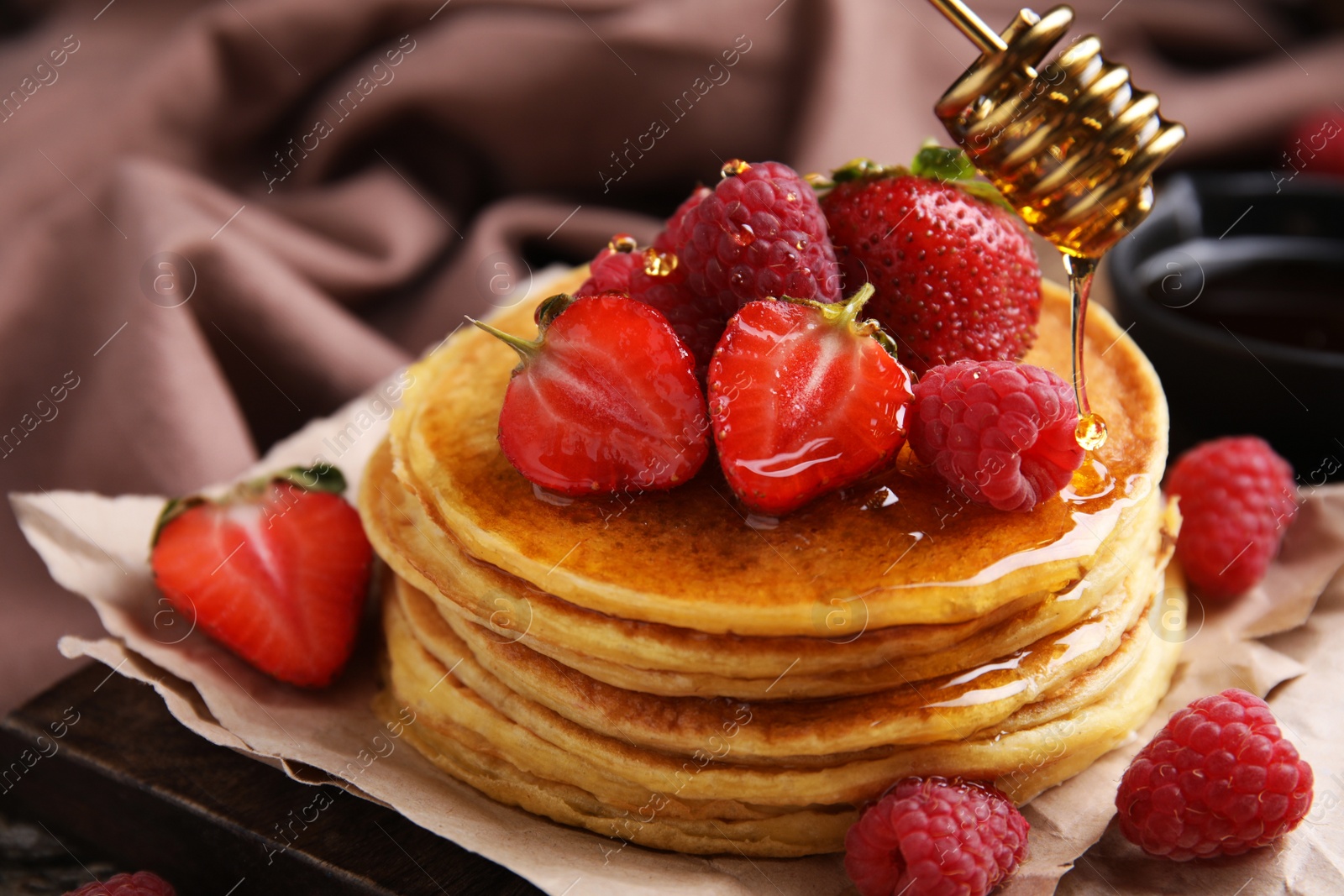 Photo of Pouring honey onto tasty pancakes with fresh berries on wooden board, closeup