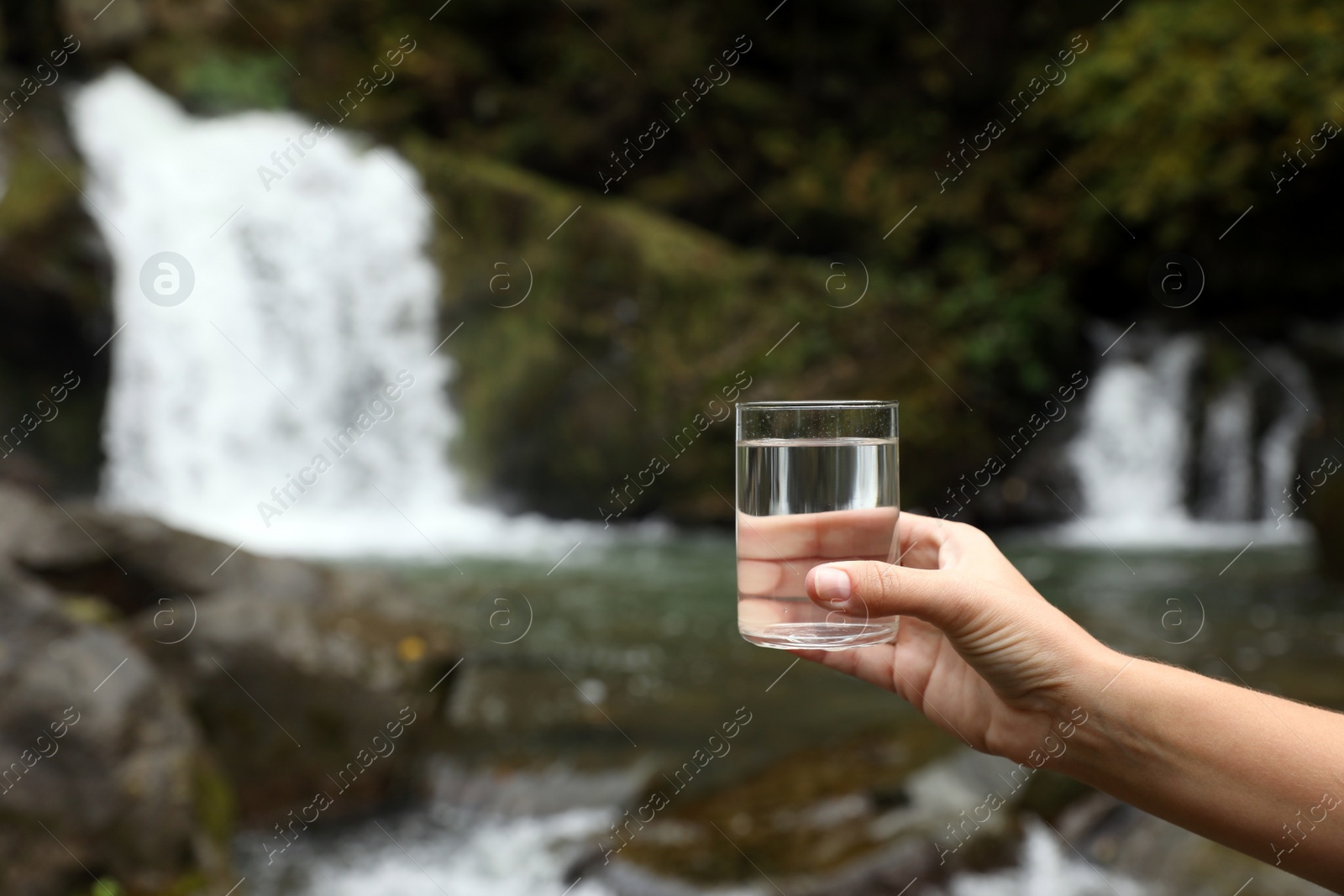 Photo of Woman holding glass of fresh water near waterfall outdoors, closeup. Space for text