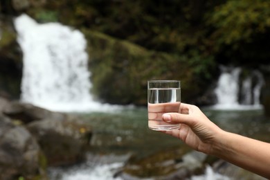Woman holding glass of fresh water near waterfall outdoors, closeup. Space for text