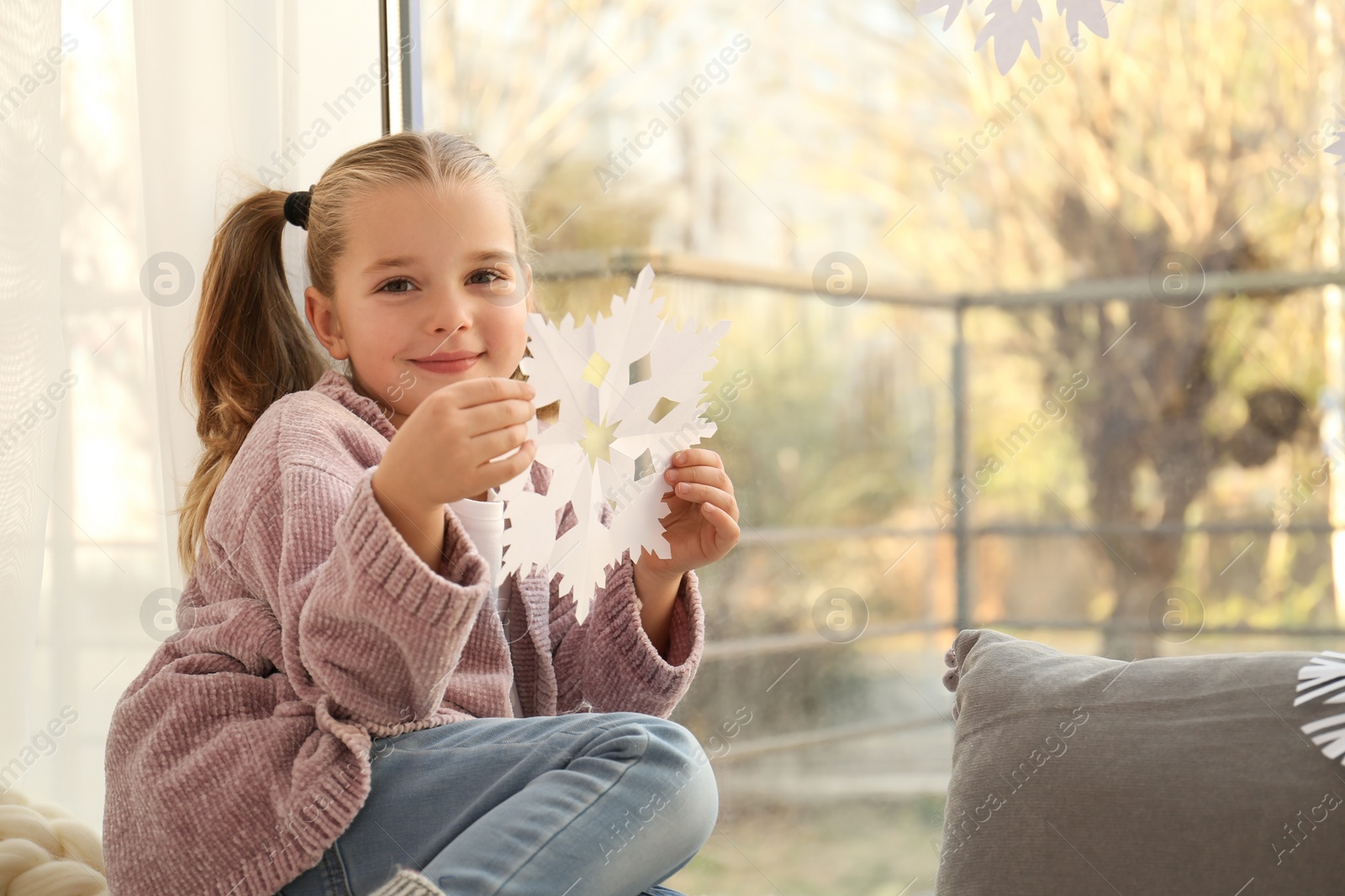 Photo of Little girl with paper snowflake near window indoors