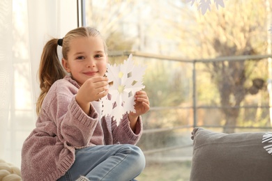 Photo of Little girl with paper snowflake near window indoors
