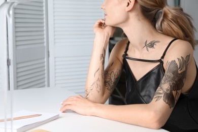 Photo of Beautiful woman with tattoos on body at table indoors, closeup