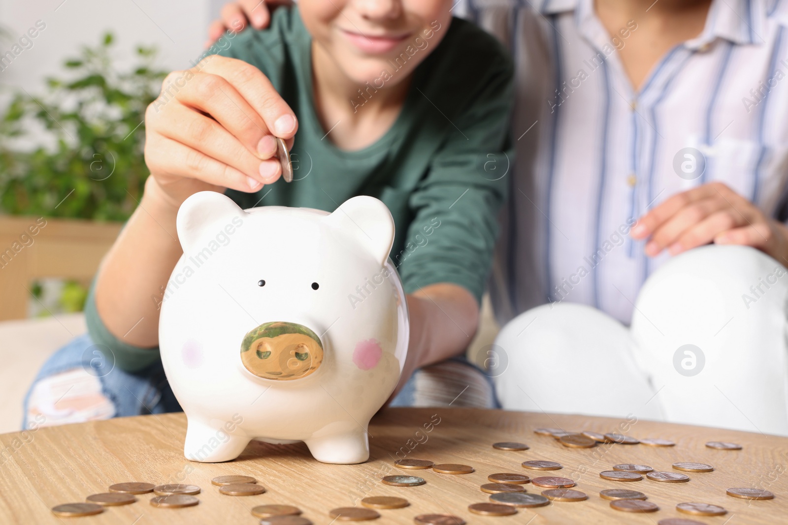 Photo of Boy with his mother putting coin into piggy bank at home, closeup