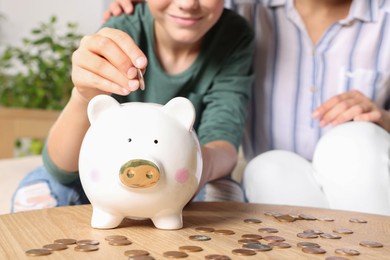Boy with his mother putting coin into piggy bank at home, closeup