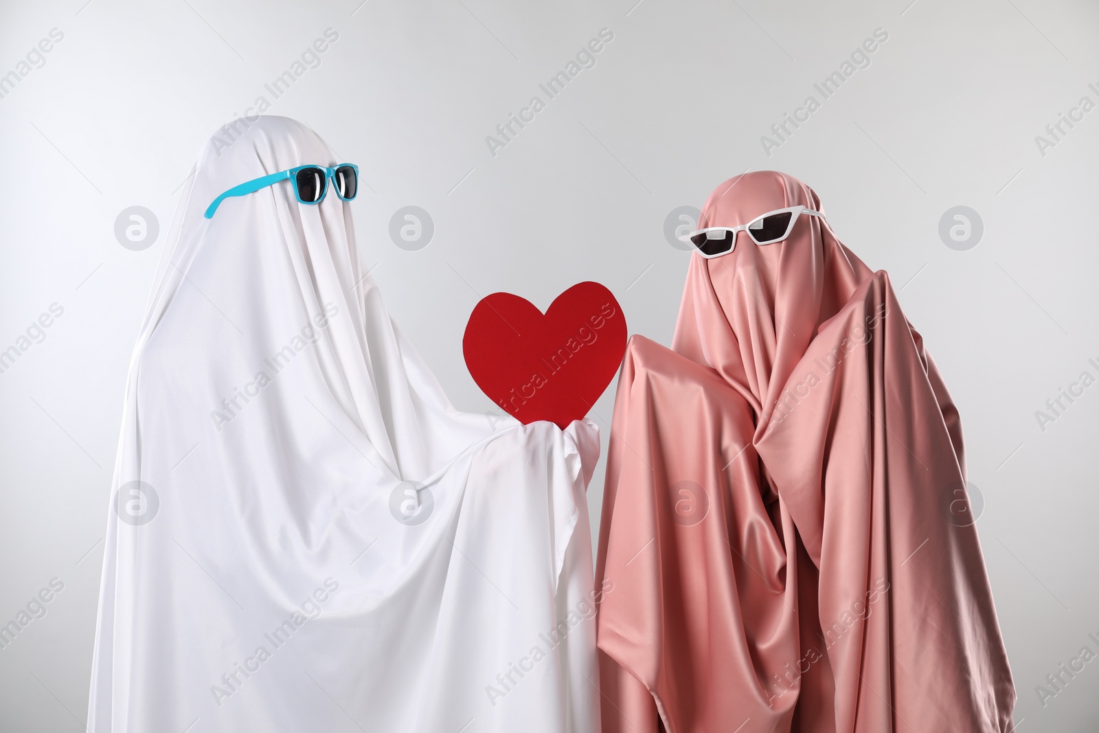 Photo of Cute ghosts. Couple in color sheets and sunglasses with red heart on light grey background