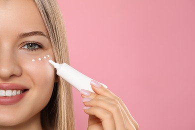 Photo of Young woman applying cream under eyes on pink background, closeup. Space for text