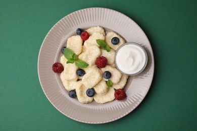 Photo of Plate of tasty lazy dumplings with berries, mint leaves and sour cream on dark green background, top view