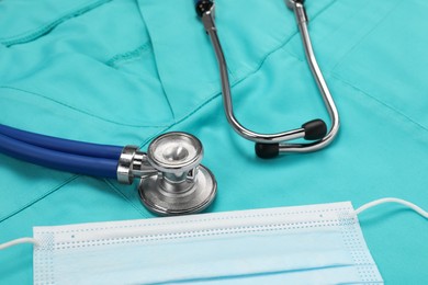 Photo of Stethoscope and protective mask on turquoise medical uniform, closeup