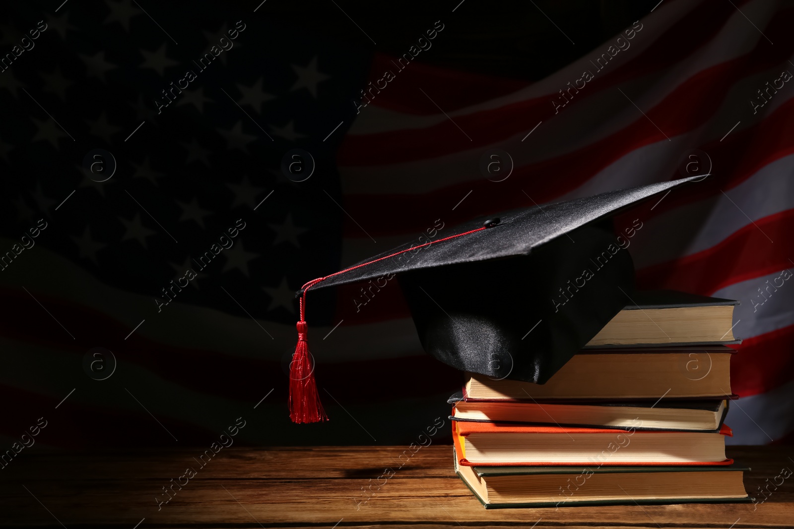 Photo of Graduation hat and books on wooden table against American flag in darkness, space for text