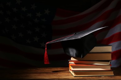 Photo of Graduation hat and books on wooden table against American flag in darkness, space for text