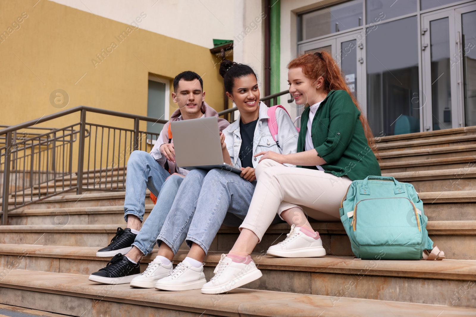 Photo of Happy young students studying together with laptop on steps outdoors