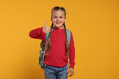Happy schoolgirl with backpack showing thumb up gesture on orange background