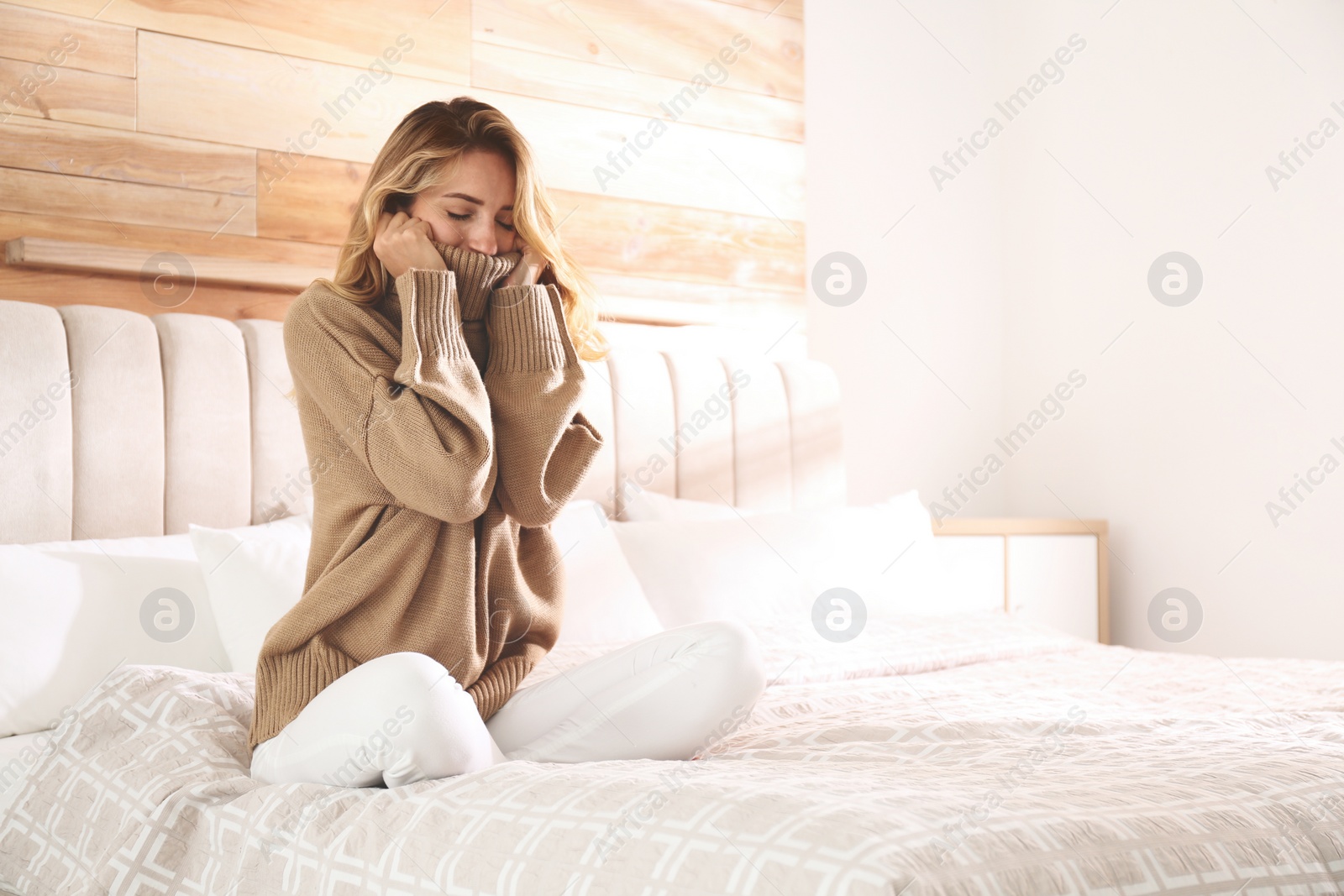 Photo of Beautiful young woman wearing warm brown sweater on bed at home. Space for text