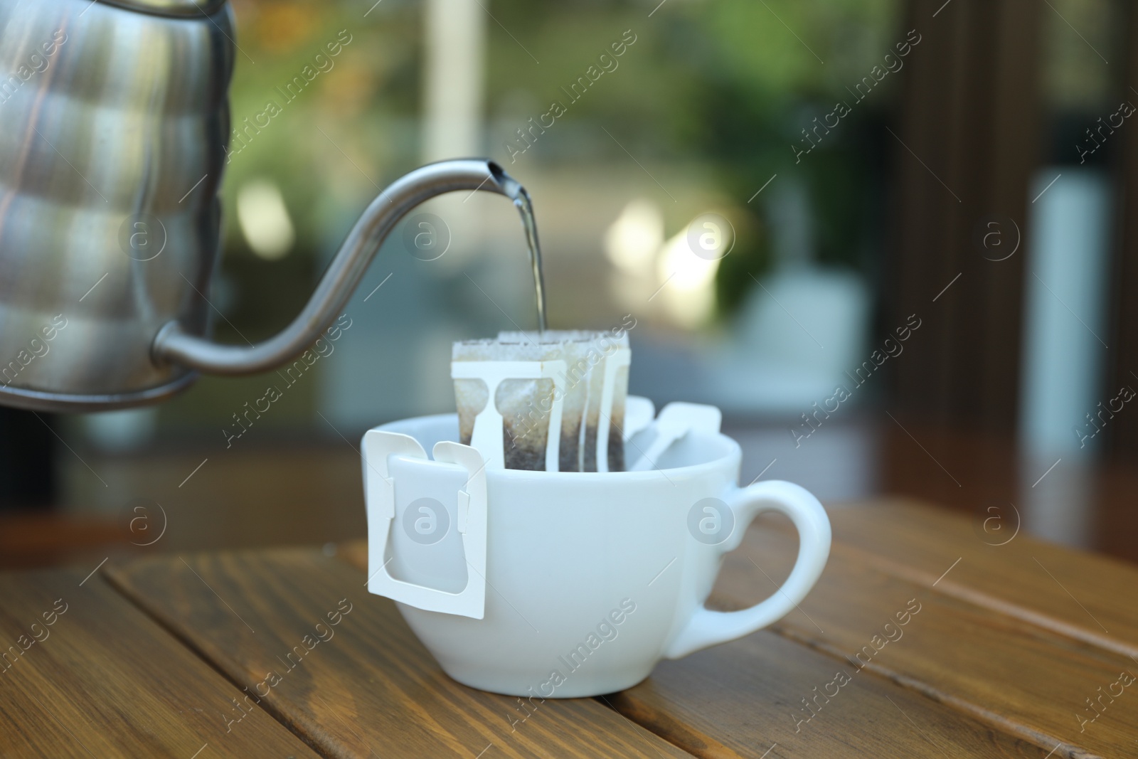 Photo of Pouring hot water into cup with drip coffee bag from kettle on wooden table, closeup