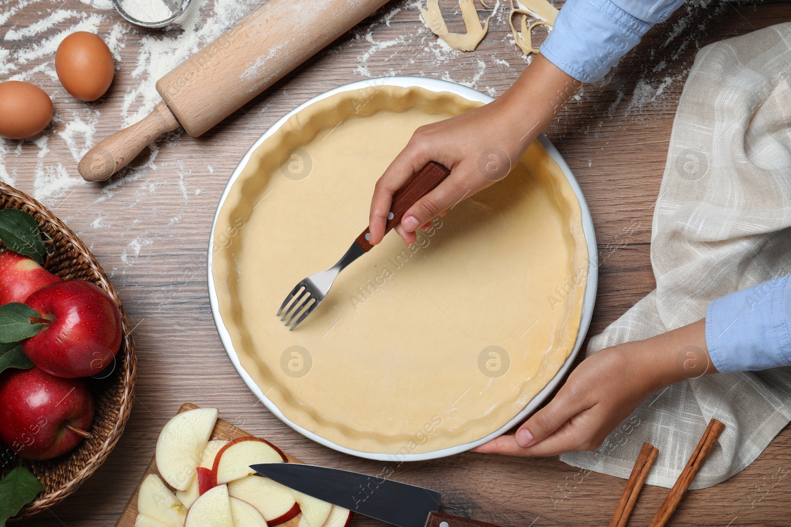 Photo of Woman making holes in raw dough with fork at wooden table, top view. Baking apple pie