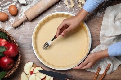 Photo of Woman making holes in raw dough with fork at wooden table, top view. Baking apple pie