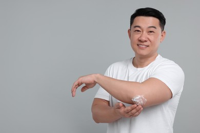Handsome man applying body cream onto his elbow on light grey background. Space for text