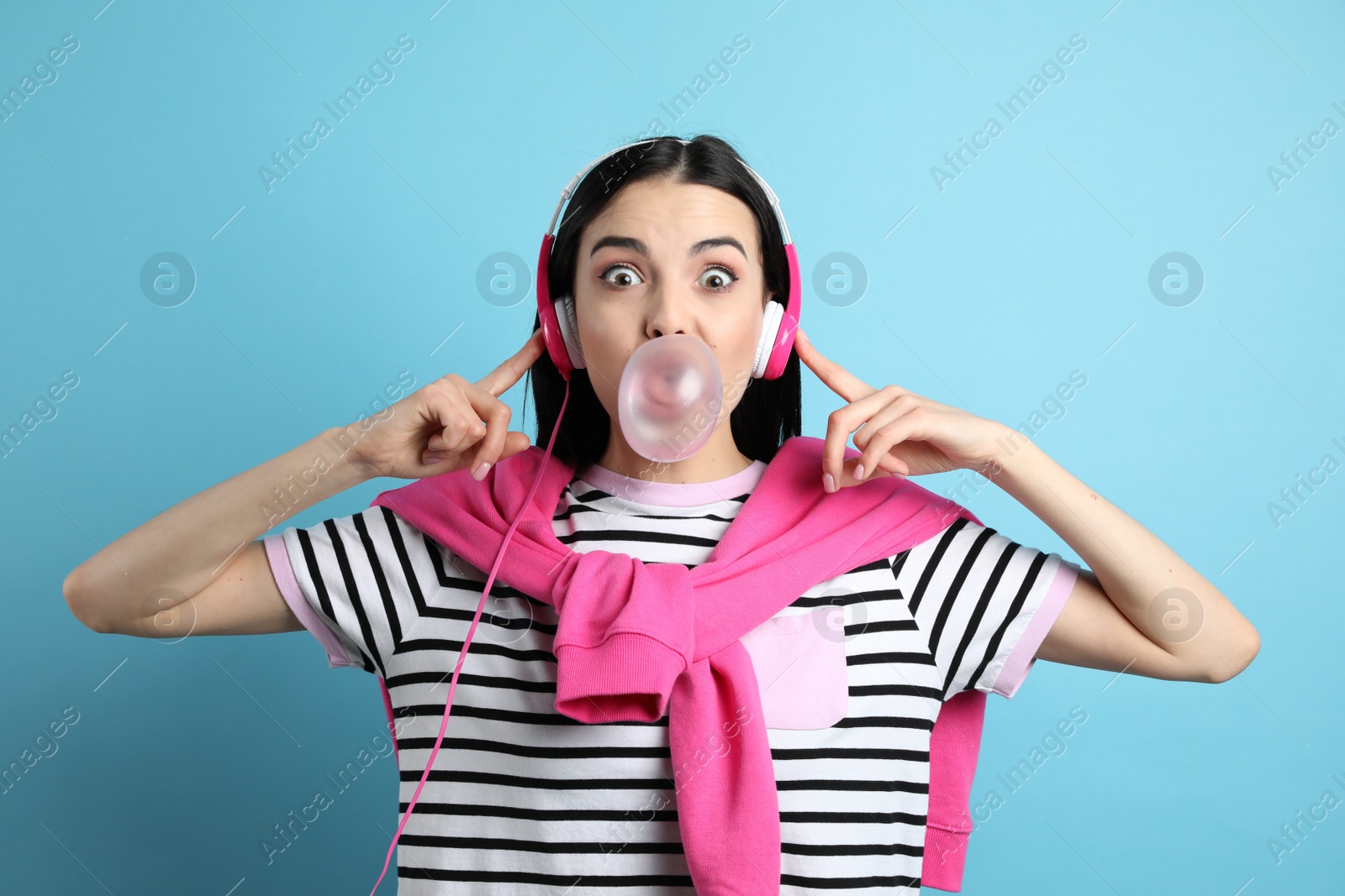 Photo of Fashionable young woman with headphones blowing bubblegum on light blue background