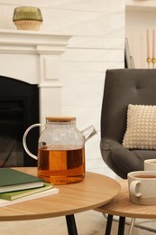 Photo of Teapot, cups of hot drink and books on wooden tables near armchair in room. Interior design
