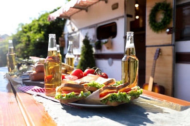 Photo of Delicious sandwiches and bottles of beer on wooden table near motorhome on sunny day. Camping season