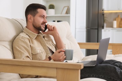 Photo of Man talking on phone while working with laptop on sofa at home