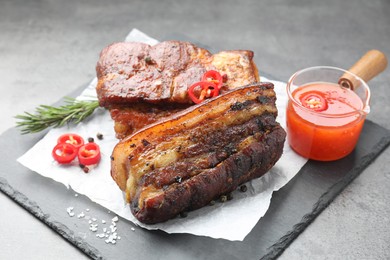 Photo of Pieces of baked pork belly served with sauce, rosemary and chili pepper on grey table