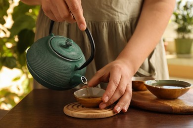 Photo of Woman pouring freshly brewed tea from teapot into cup at wooden table indoors, closeup. Traditional ceremony