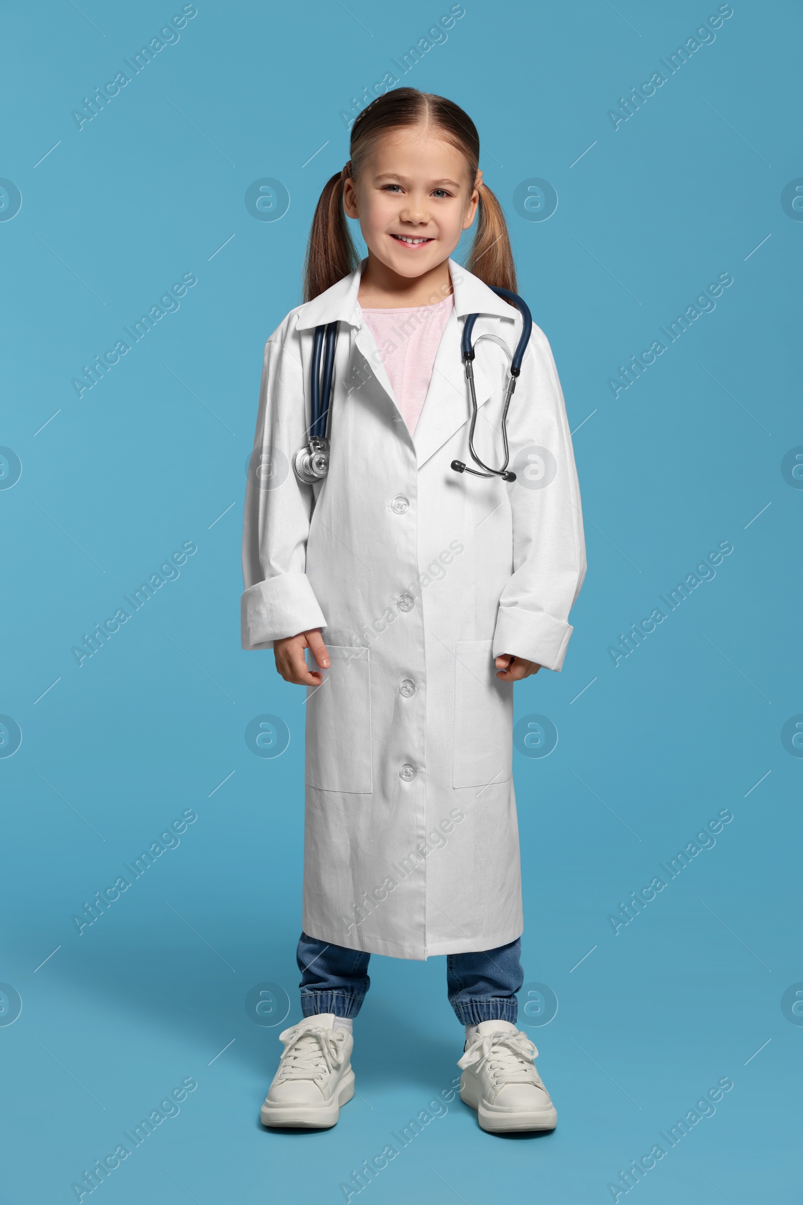 Photo of Little girl in medical uniform with stethoscope on light blue background