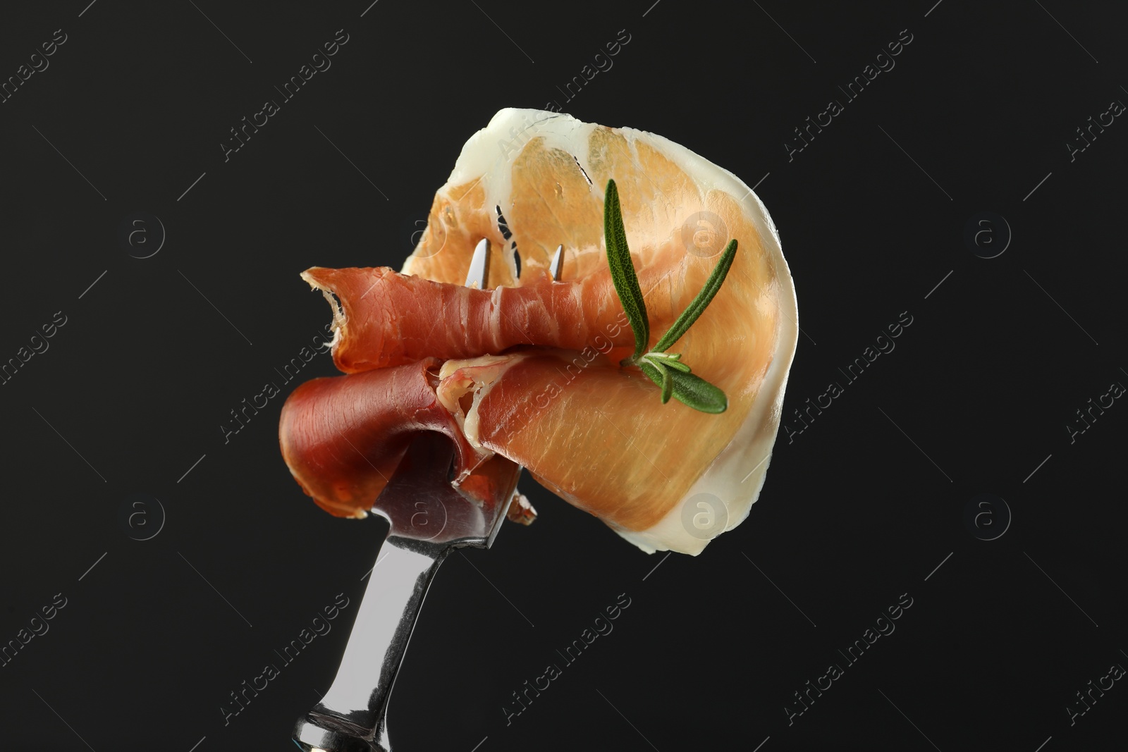 Photo of Fork with slice of tasty jamon and rosemary on black background, closeup