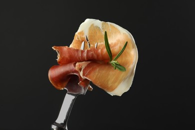 Photo of Fork with slice of tasty jamon and rosemary on black background, closeup