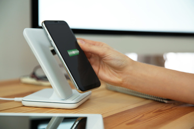 Photo of Woman putting mobile phone onto wireless charger at wooden table, closeup. Modern workplace accessory