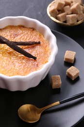 Photo of Delicious creme brulee in bowl, vanilla pods, sugar cubes spoon on dark gray table, closeup