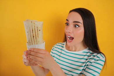 Emotional young woman holding tasty shawarma on yellow background