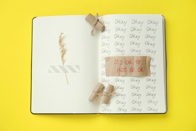 Photo of Phrase It`s Ok to Not Be Ok, rolled pieces of paper and dry flower attached with adhesive tape in notebook on yellow background, top view