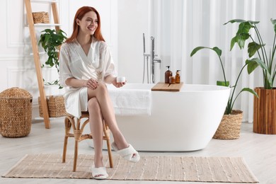 Beautiful young woman applying body cream onto legs in bathroom, space for text