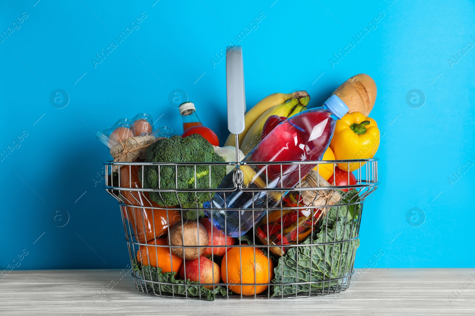 Photo of Shopping basket with grocery products on white wooden table against light blue background