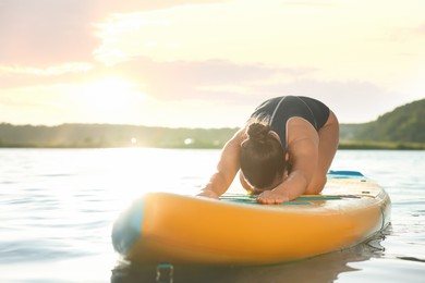 Young woman practicing yoga on color SUP board on river at sunset