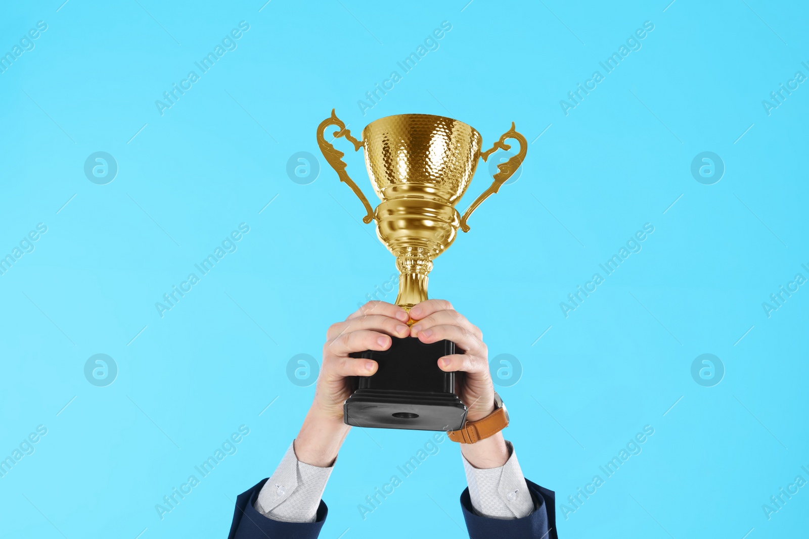 Photo of Businessman holding gold trophy cup on light blue background, closeup