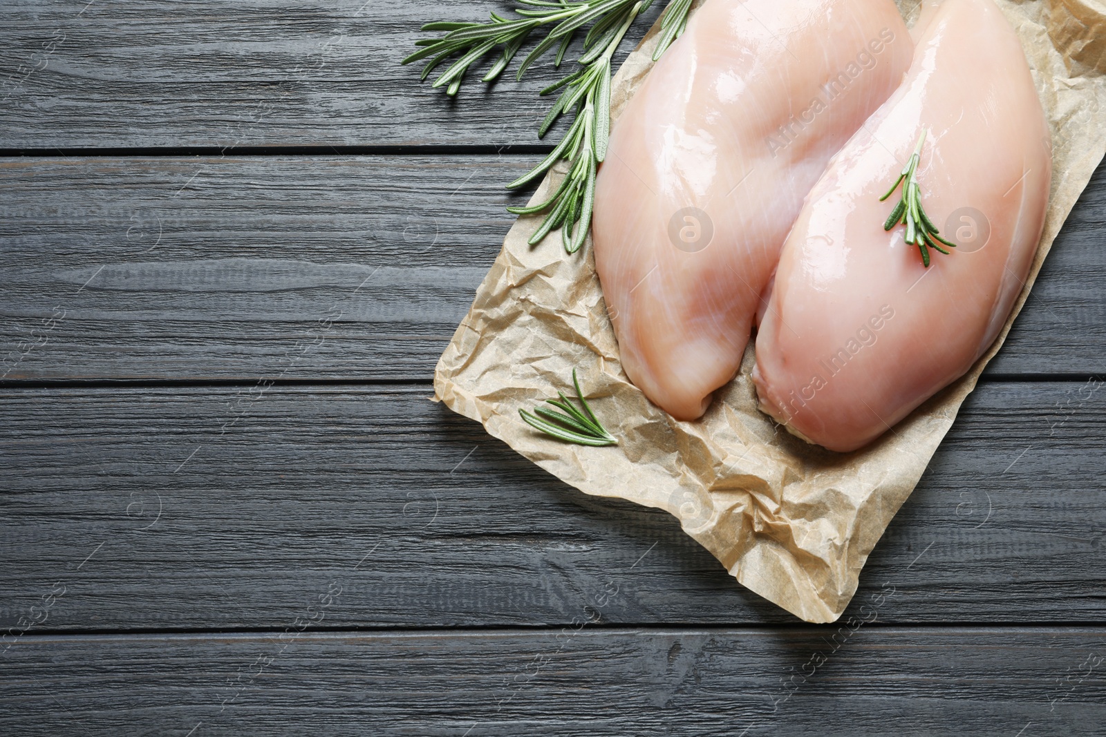 Photo of Parchment with raw chicken breasts and rosemary on wooden background, top view. Space for text