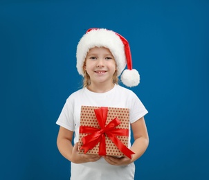 Photo of Cute little child wearing Santa hat with Christmas gift on blue background