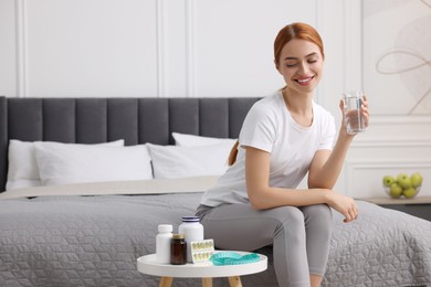 Happy young woman holding glass of water near table with pills and measuring tape in room, space for text. Weight loss