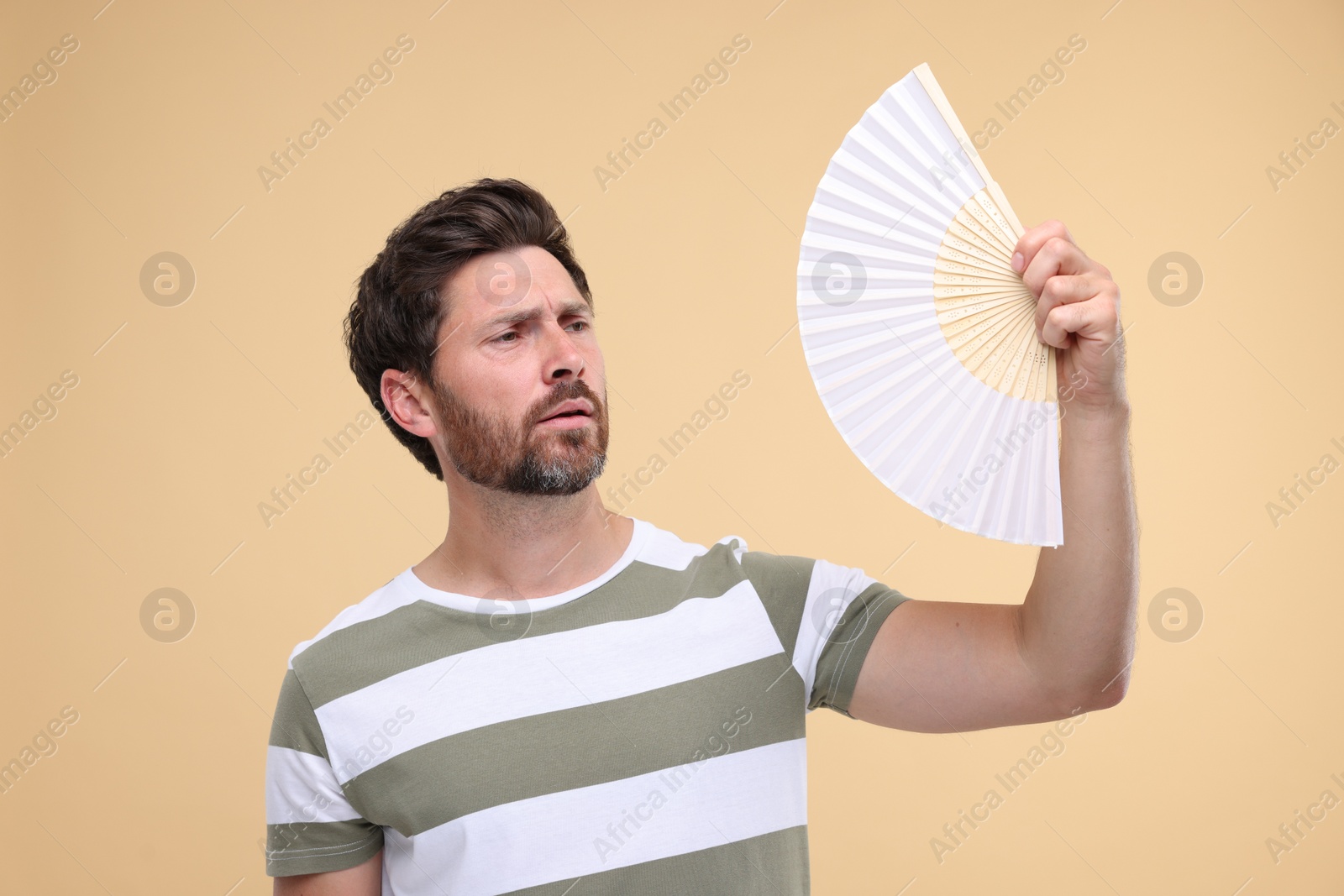 Photo of Unhappy man with hand fan suffering from heat on beige background