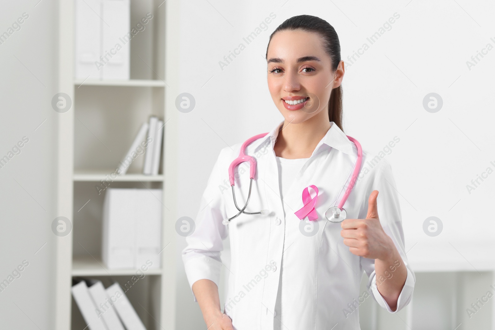 Photo of Mammologist with pink ribbon showing thumbs up in hospital, space for text. Breast cancer awareness