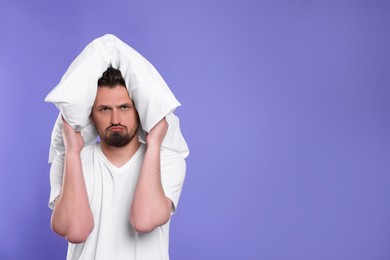 Photo of Unhappy man covering ears with pillow on violet background, space for text. Insomnia problem
