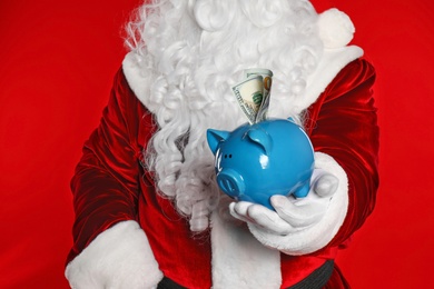 Santa Claus holding piggy bank with dollar banknotes on red background, closeup