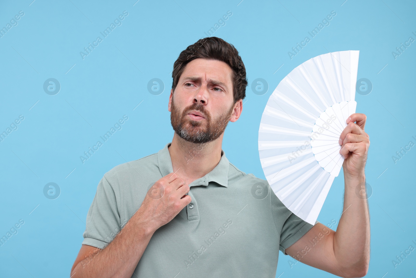 Photo of Unhappy man with hand fan suffering from heat on light blue background