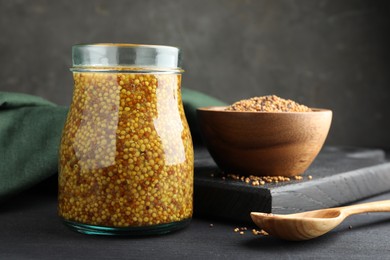 Photo of Whole grain mustard in jar and dry seeds on black wooden table