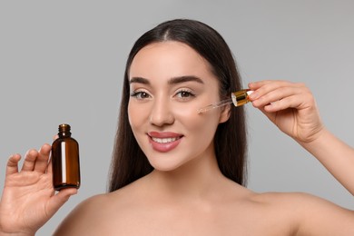 Happy young woman with bottle applying essential oil onto face on light grey background