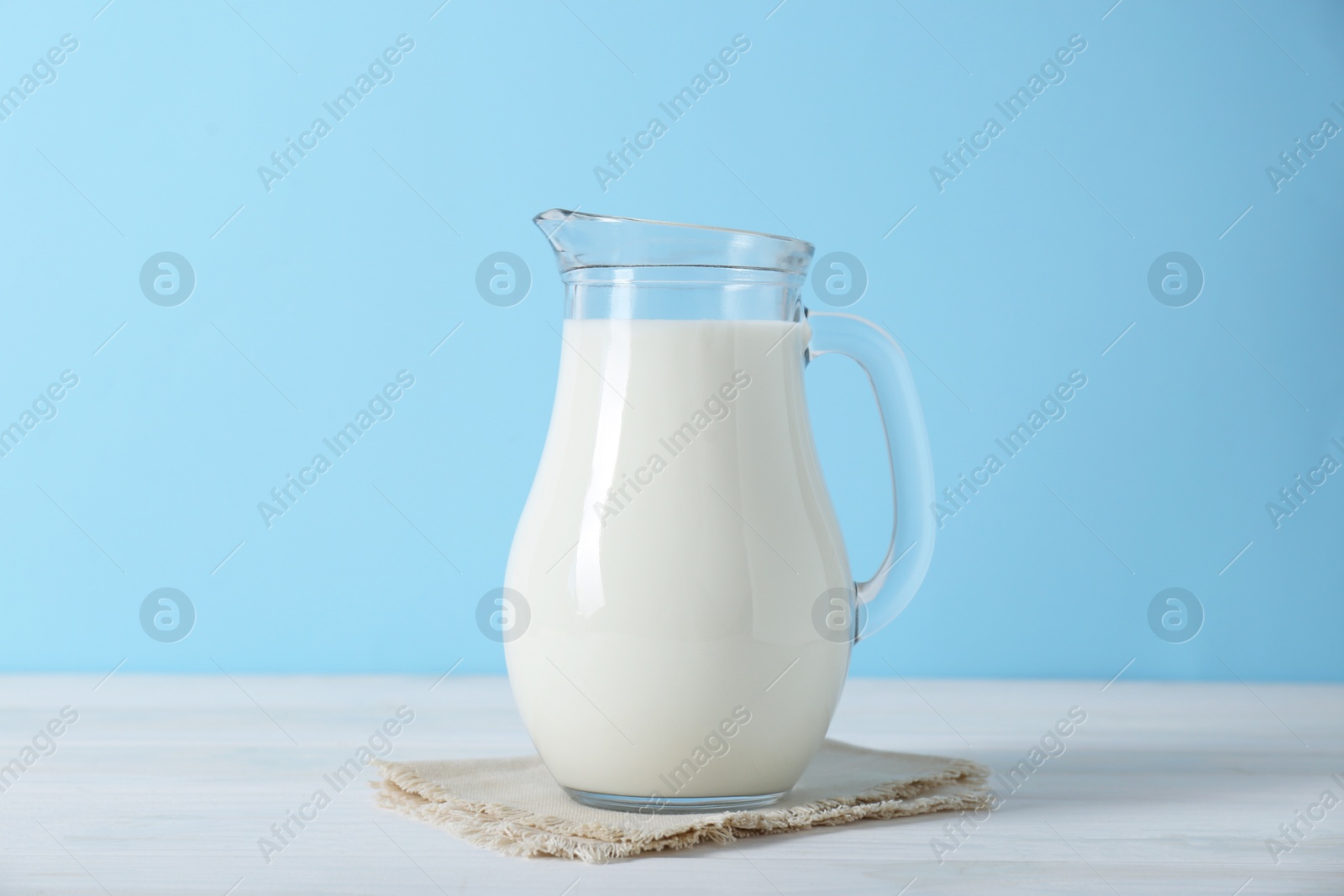 Photo of Jug of fresh milk on white wooden table against light blue background