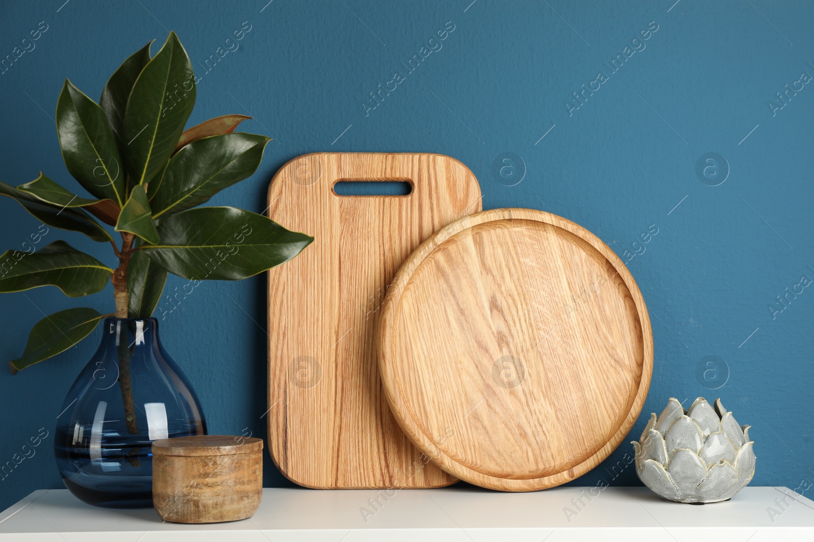 Photo of Wooden cutting boards, branch with green leaves and decor on white table near blue wall